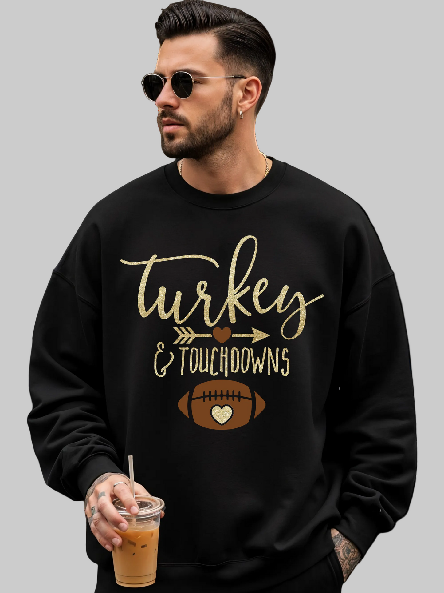 A man wearing a black crewneck sweatshirt with a gold and brown 'Turkey & Touchdowns' football graphic, holding an iced coffee against a plain white background.