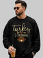 A man wearing a black crewneck sweatshirt with a gold and brown 'Turkey & Touchdowns' football graphic, holding an iced coffee against a plain white background.