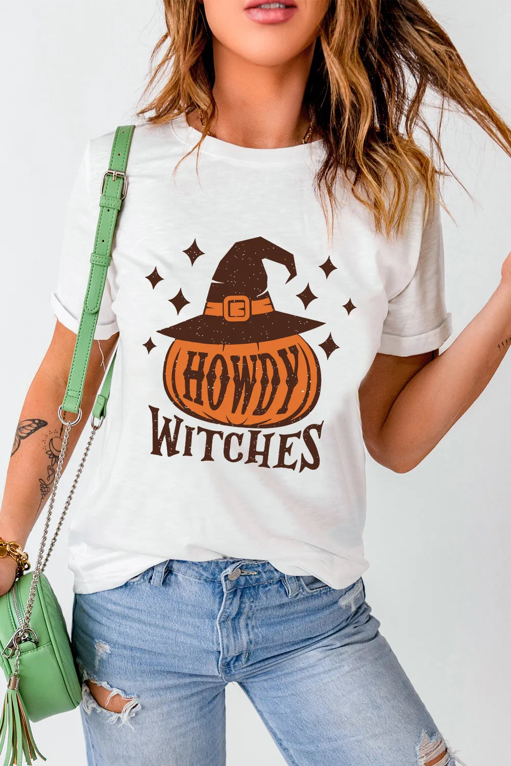 Woman wearing white HOWDY WITCHES round neck short sleeve t-shirt with pumpkin and witch hat graphic, paired with ripped jeans and green purse.